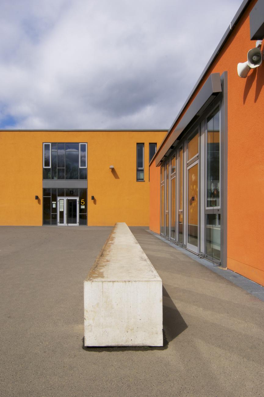 Schulhof mit Blick auf das Forum und eines der beiden Unterrichtsgebäude. Schulhof mit Blick auf eine langestreckte Betonbank vor einem eingeschossigen Unterrichtsgebäude mit Blick auf das sich links anschließende, eingeschossige Forum.